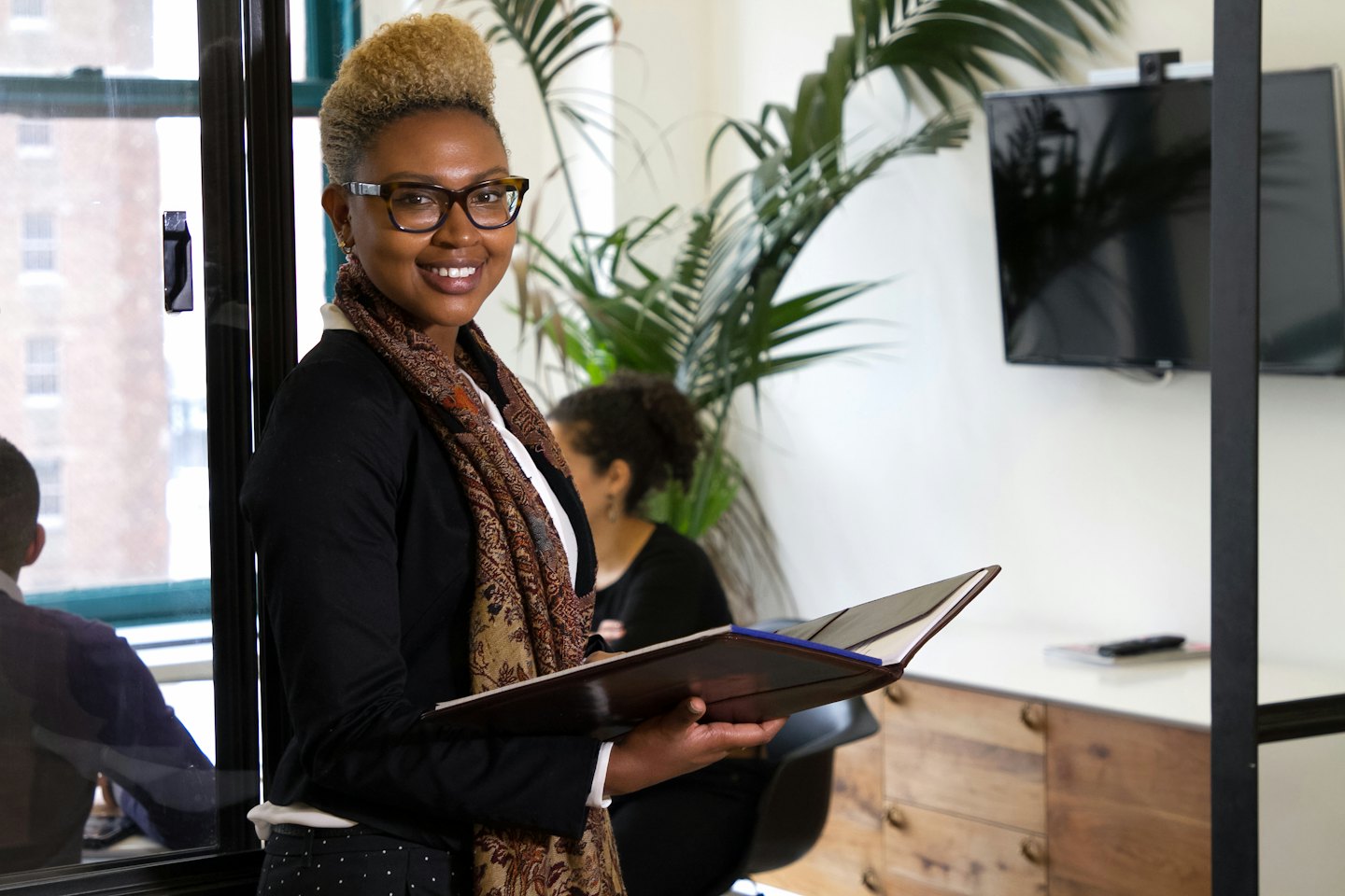 A Black woman in a modern office holds an open folder and smiles.